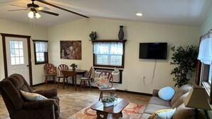 Ein Wohnzimmer mit Deckenventilator, einem Essbereich mit Tisch und Stühlen, einem an der Wand befestigten Fernseher und einem großen Fenster mit Vorhängen.
