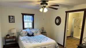 Ein Schlafzimmer mit Bett, Nachttisch, Fenster mit Vorhängen, Deckenventilator, einer Uhr an der Wand und einem durchgegangenen Tür erreichbaren Badezimmer.