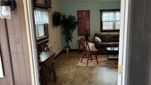 Ein offenes Durchgangsbild zu einem Wohnzimmer mit Holzboden, einer Couch, einem Sessel, einem Tisch und einem Blumentopf.