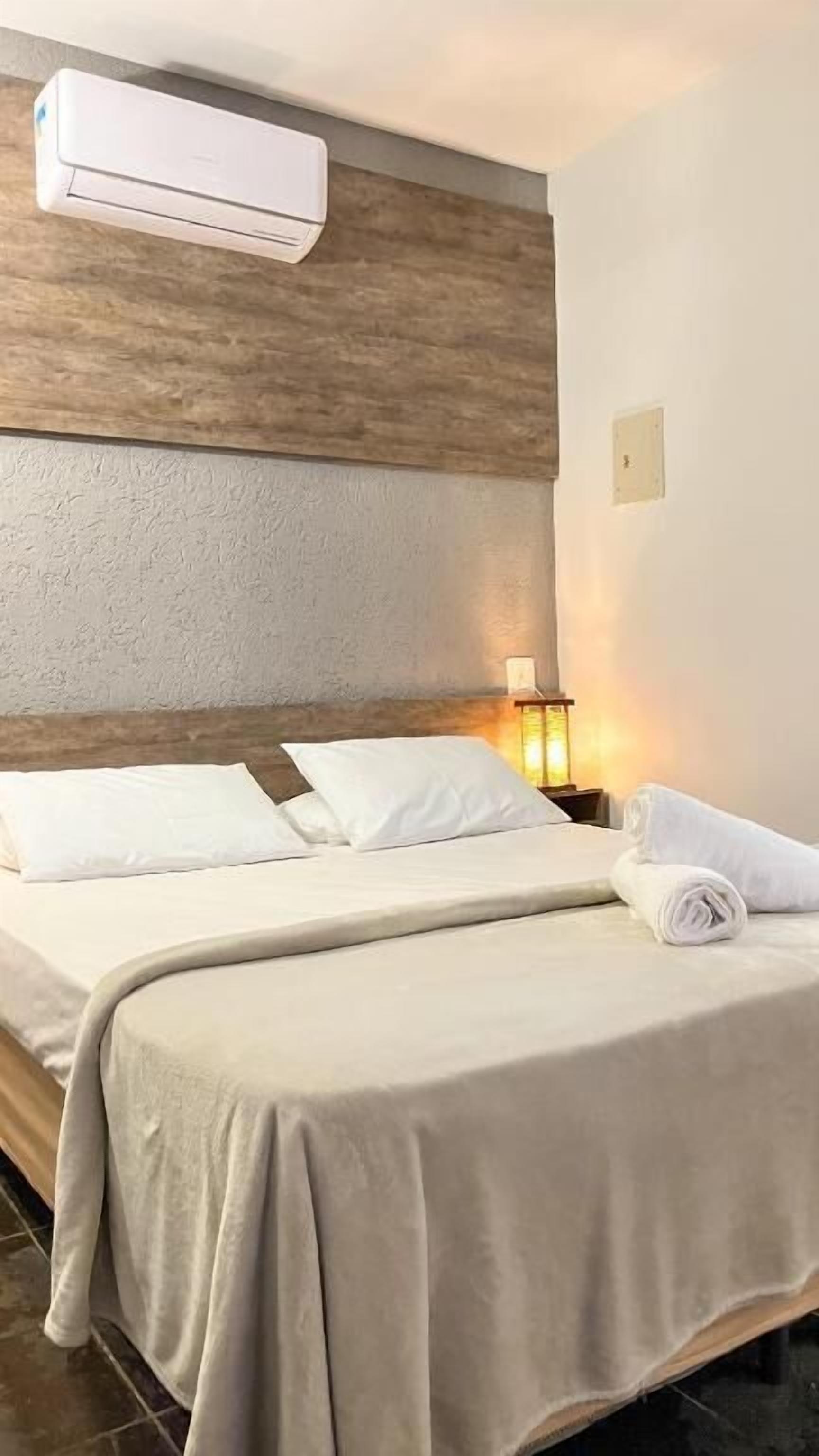 A hotel room with a bed, a nightstand, a lamp, and an air conditioning unit.