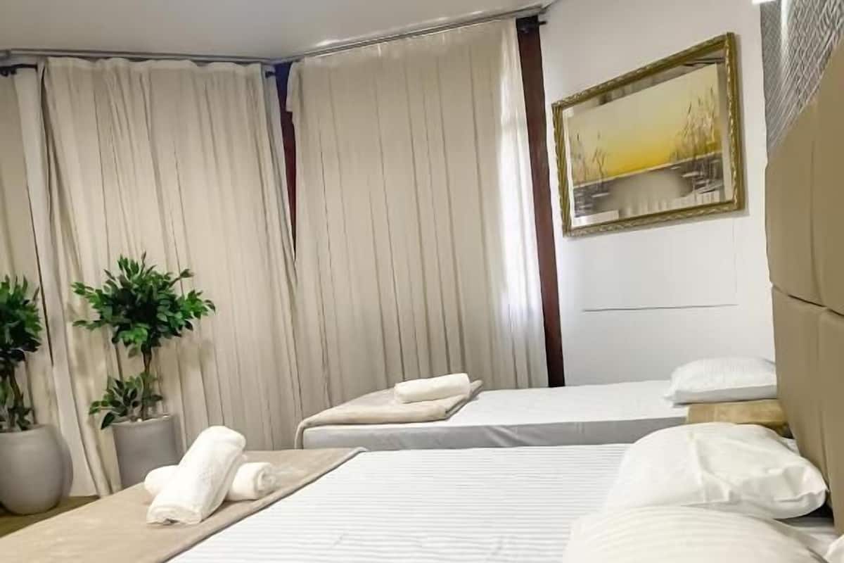 A hotel room with two beds, a large window with curtains, a painting on the wall, and a potted plant.