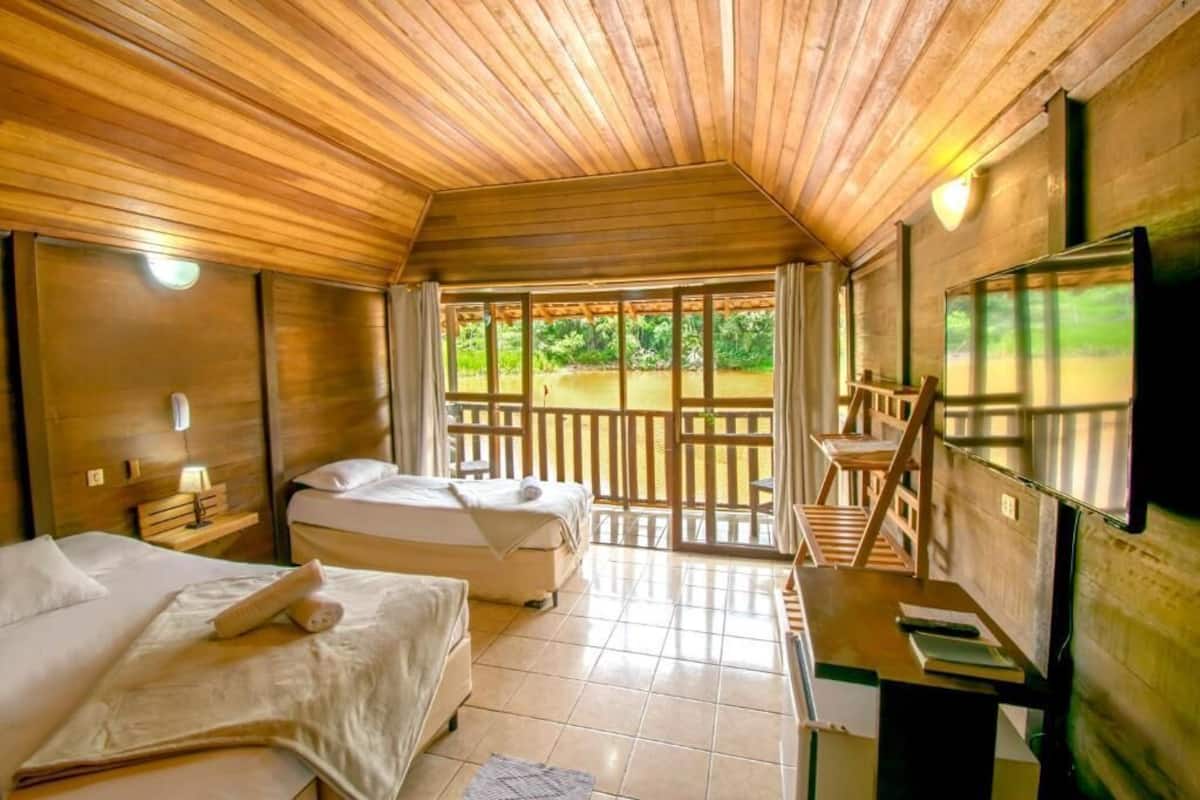 A room with two beds, a desk, a television, and a balcony with a view of greenery.