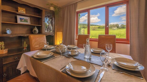 A dining area with a table set for four, a wooden bookshelf, and a window with a view of a green field.