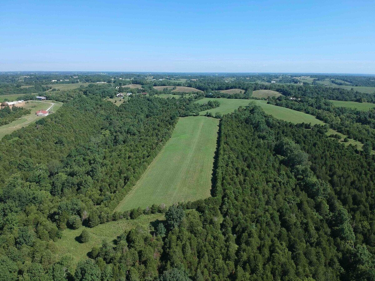 An aerial view of a rural landscape with a large green field, dense forested areas, and scattered buildings.