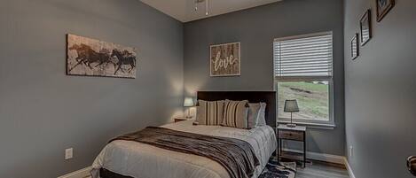 A bedroom with a bed, a ceiling fan, a window with blinds, and artwork on the walls.