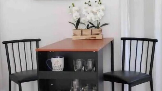 Un comedor con una mesa de madera y dos sillas negras, una estantería negra con diversos objetos, y orquídeas blancas sobre la mesa.