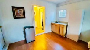 Una cocina compacta con refrigerador, un lavamanos y gabinetes de madera. Una puerta da a un pasillo con una fuente de luz.
