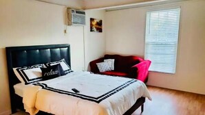 Un dormitorio con cama, sofá rojo, ventana con persianas y un equipo de aire acondicionado.