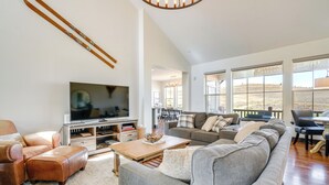 A spacious living room with a large gray sectional sofa, a wooden coffee table, and a flat-screen TV on a wooden stand. There are large windows offering a view of the outdoors.
