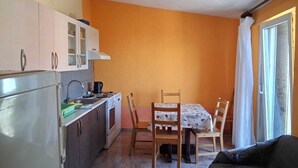 Eine kompakte Küche mit Herd, Spüle und Schränken, ein Esstisch mit Stühlen und ein Sofa in einem Raum mit orangefarbenen Wänden und einem fenster mit Vorhängen.
