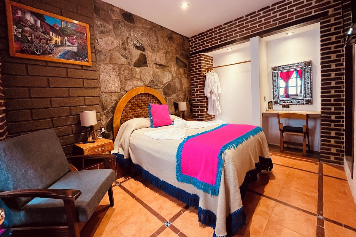 Ein Schlafzimmer mit Steinwand, einem Bett mit blauer und weißer Bettdecke, einem Holz-Kopfteil, einem Stuhl, einem Nachttisch und einem Bild an der Wand.
