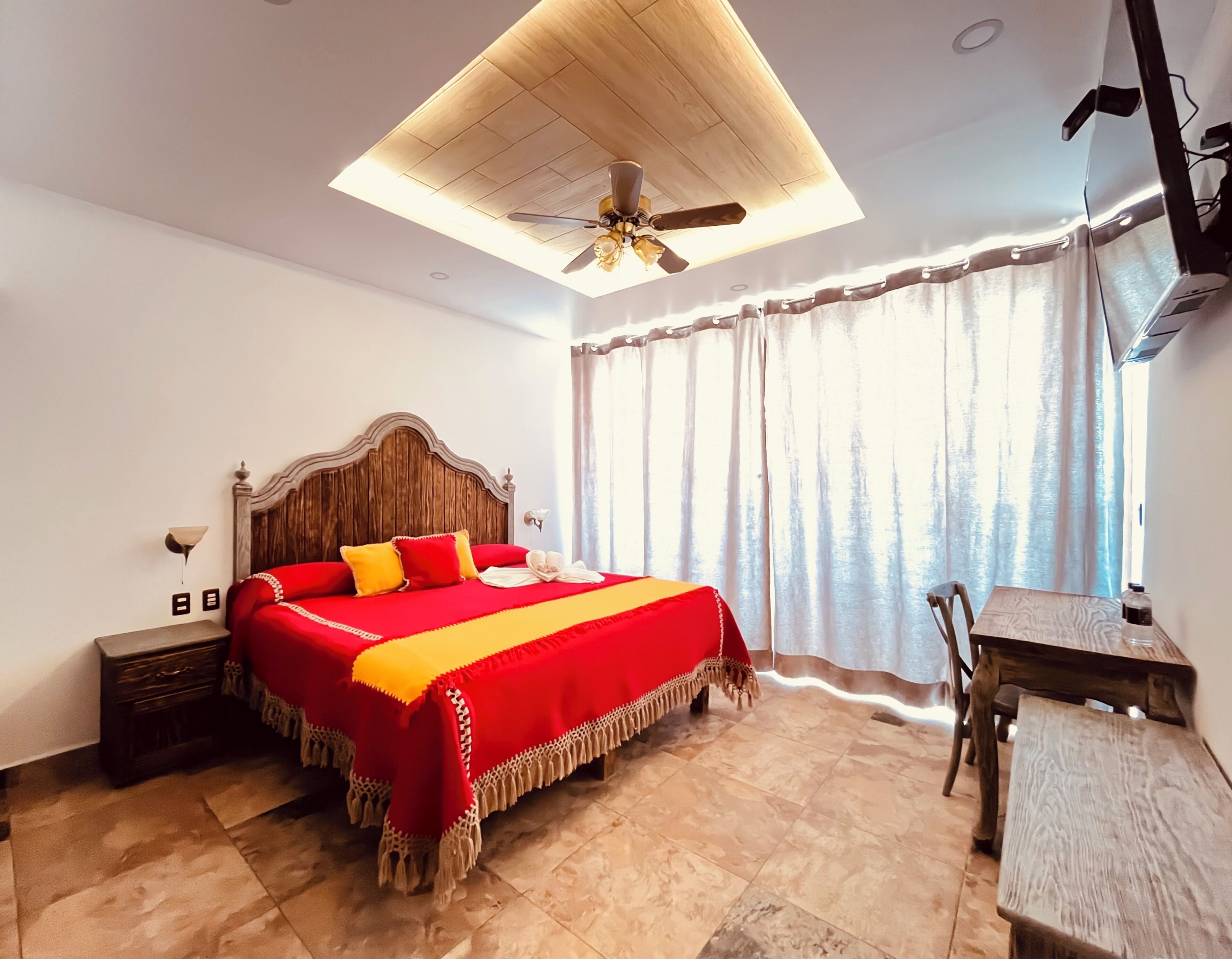 A bedroom with a large bed, a wooden ceiling fan, a desk, and a window with curtains.