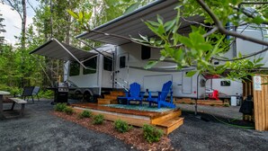 A recreational vehicle with a covered porch, a grill, and blue chairs. - RV 5   Rustic Charm w/ Queen Bed with Forest Background (Kenora)