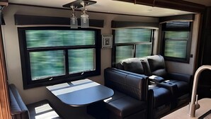 A compact RV interior with a dining area, seating, and large windows. - RV 5   Rustic Charm w/ Queen Bed with Forest Background (Kenora)