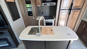 A modern kitchen with a central island, stainless steel appliances, and wooden cabinetry. - RV 5   Rustic Charm w/ Queen Bed with Forest Background (Kenora)