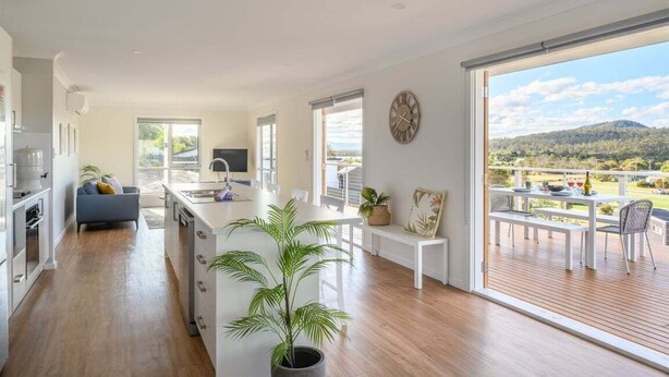 A modern kitchen with a central island, a dining area with a view, and a living space with a sofa and TV. - Ambience - Freycinet Holiday Houses (Coles Bay)