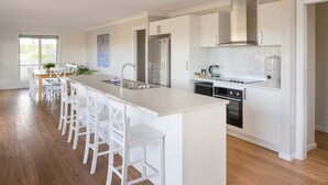 A modern kitchen with a central island, white cabinetry, stainless steel appliances, and a dining area with white chairs. - Ambience - Freycinet Holiday Houses (Coles Bay)