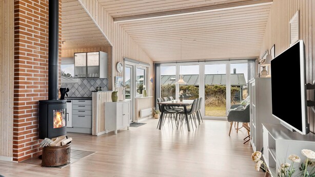 A modern kitchen with a wood-burning stove, a dining area with a table and chairs, and a large TV on the right. - Lovely home in Thisted with kitchen (Thisted)
