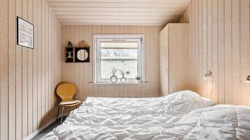 Une chambre avec un plafond en bois à lattes, un lit simple, une chaise en bois, une fenêtre donnant sur un immeuble et une étagère avec des objets décoratifs.