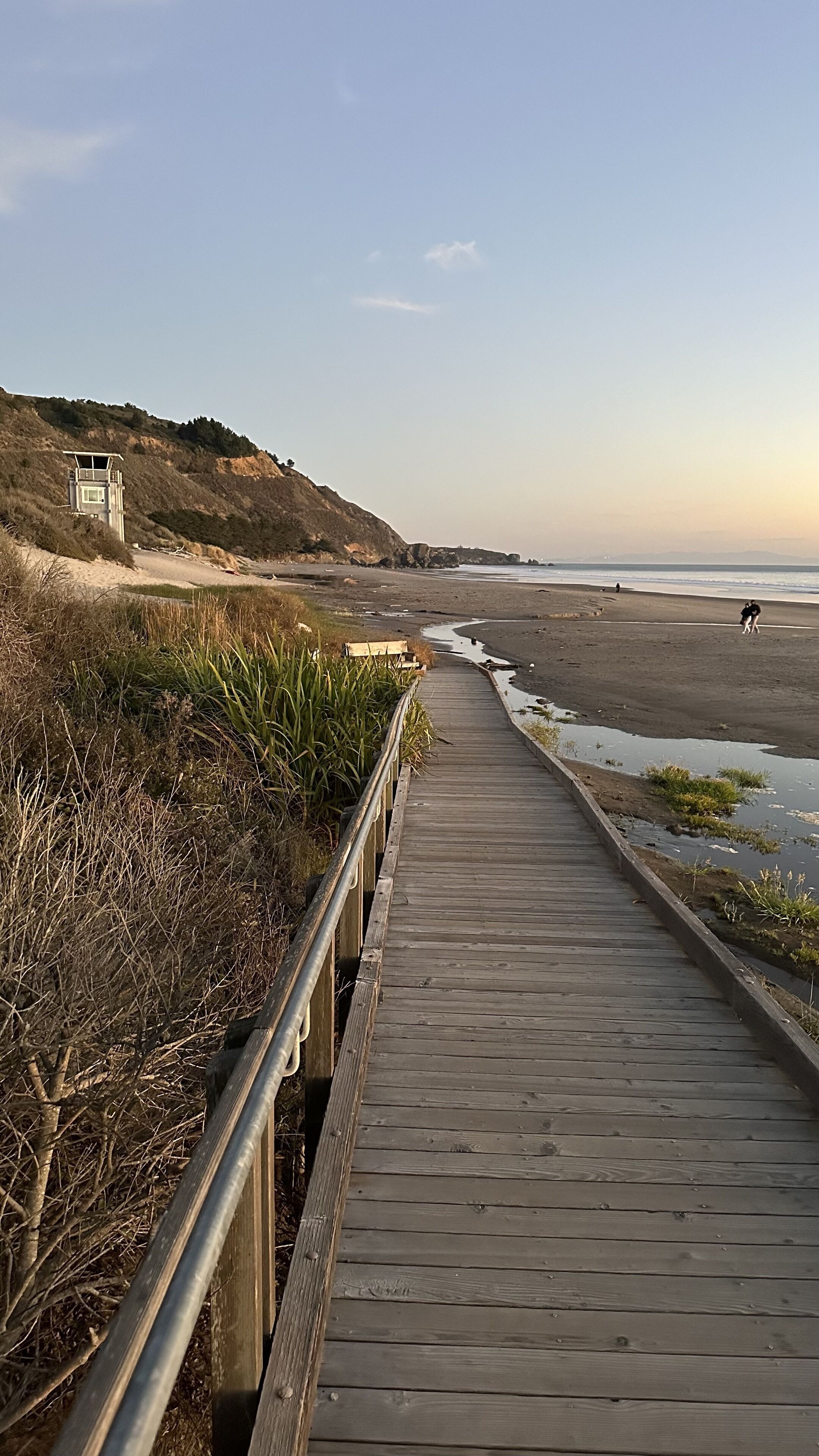 A wooden boardwalk leading to a beach with a lifeguard tower on a hillside.