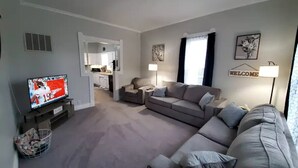 A modern living room with a grey sofa set, a TV on a stand, and a basket of newspapers. - Home Away From Home

 (Warrensburg)