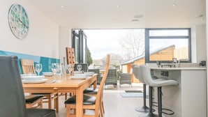 A modern dining area with a wooden table set for four, bar stools, and a large window offering a view of the outdoors. - Sailors (Falmouth)
