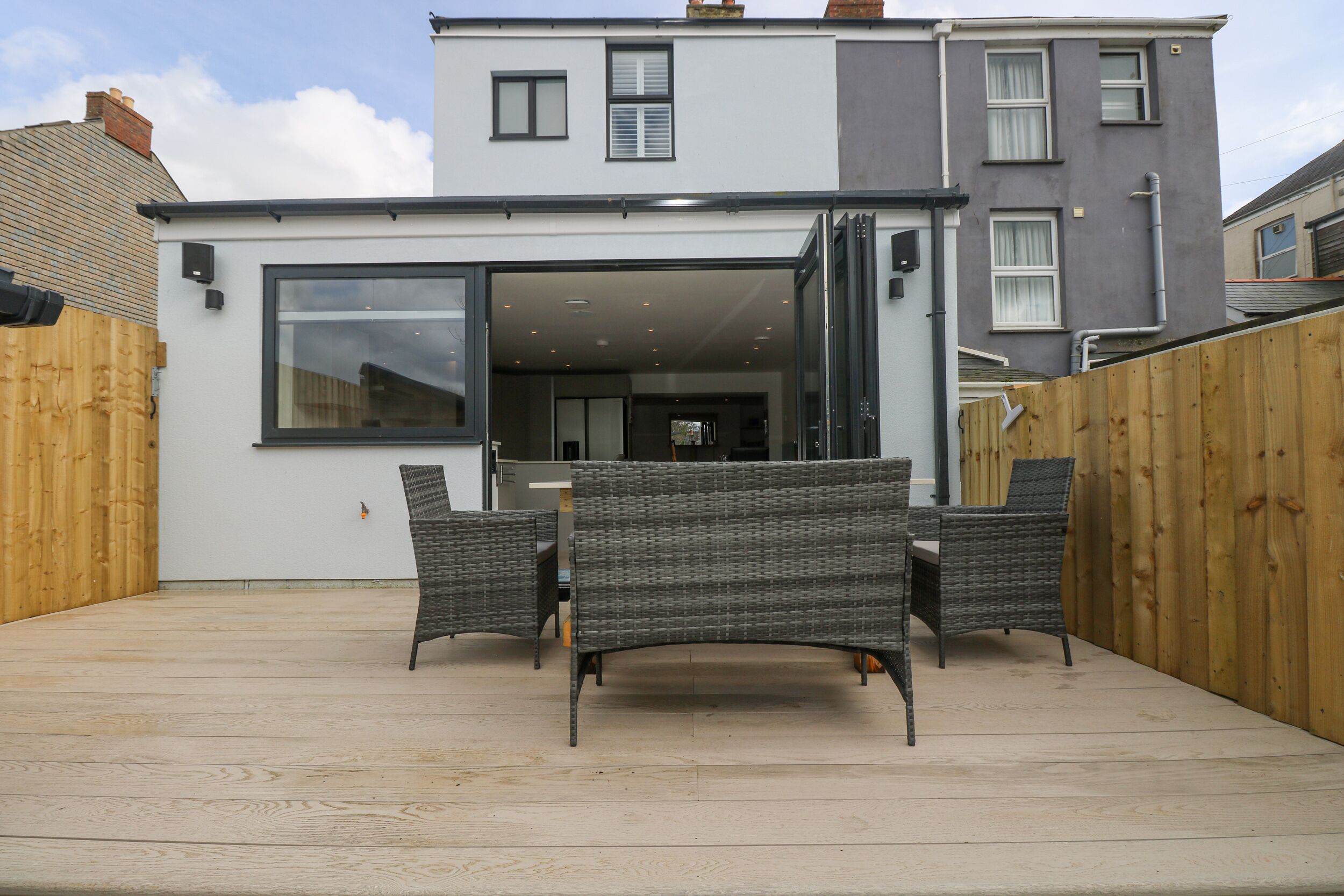 A modern patio with wicker furniture, a glass door leading to a covered area, and a wooden fence.