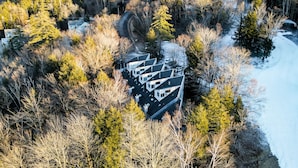 A modern, multi-story building with a unique angular design, surrounded by dense forest and snow-covered ground. - Lift Haus - New Trailside 2br Condo at Attitash - Clean • Modern • Easy (Bartlett)