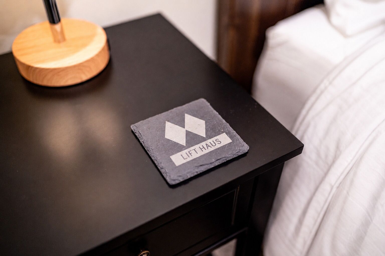 A black bedside table with a wooden lamp and a slate coaster with 'LIFT HAUS' text.