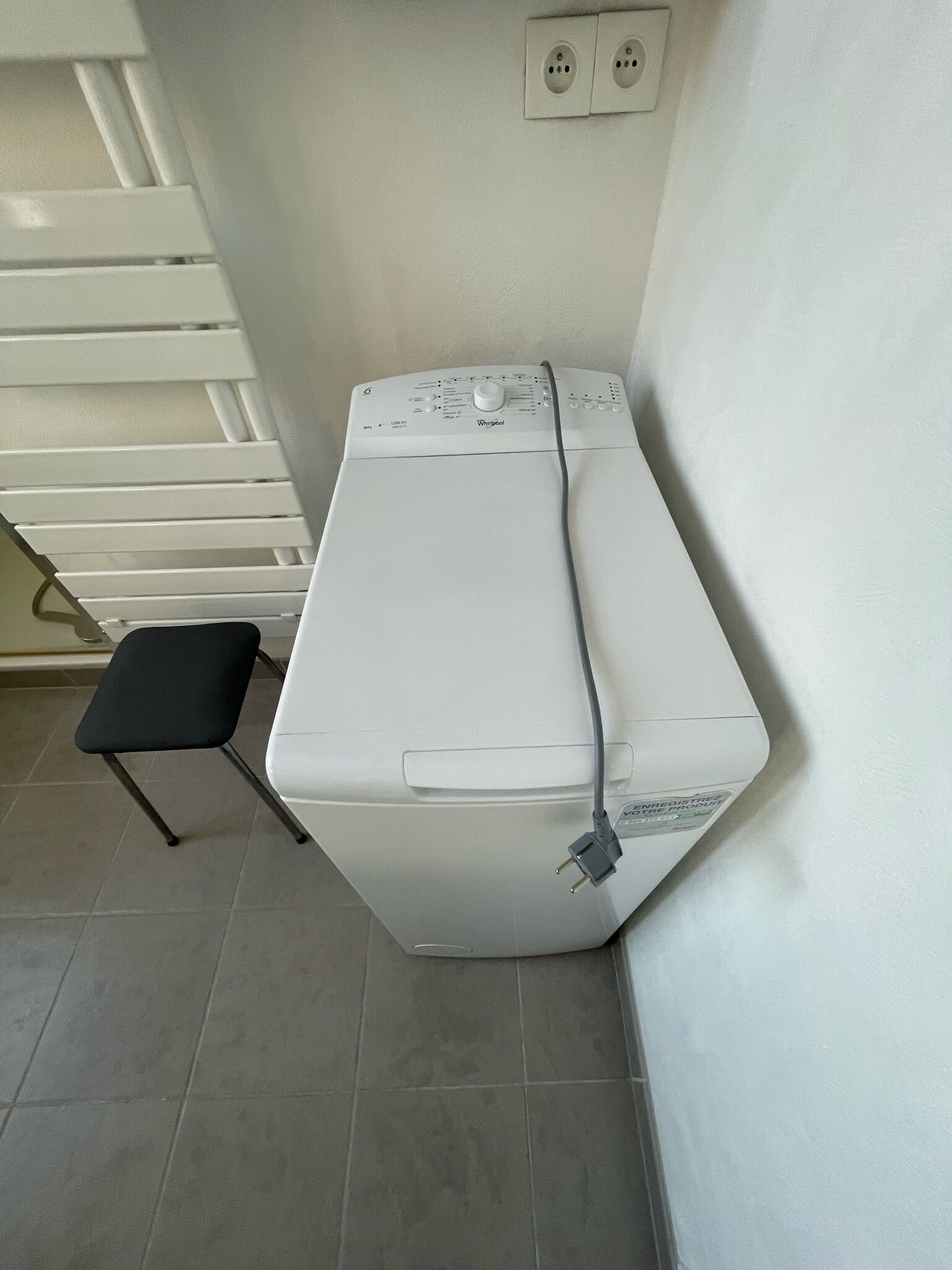 Une machine à laver blanche, dotée d’un panneau de commande et d’un câble d’alimentation branché, est placée contre un mur, juste en dessous d’une prise de courant.
