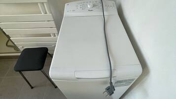 Une machine à laver blanche, dotée d’un panneau de commande et d’un câble d’alimentation branché, est placée contre un mur, juste en dessous d’une prise de courant.