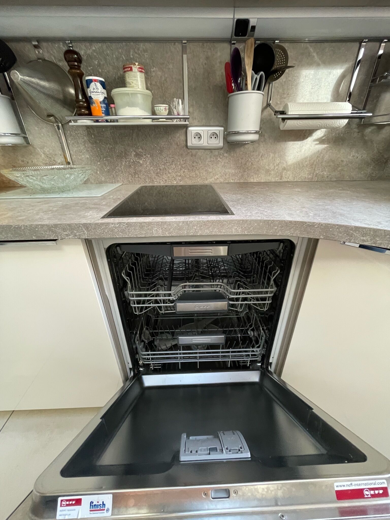 Una cocina con lavavajillas integrado, un lavamanos de acero inoxidable y una encimera con diversos utensilios y electrodomésticos de cocina.