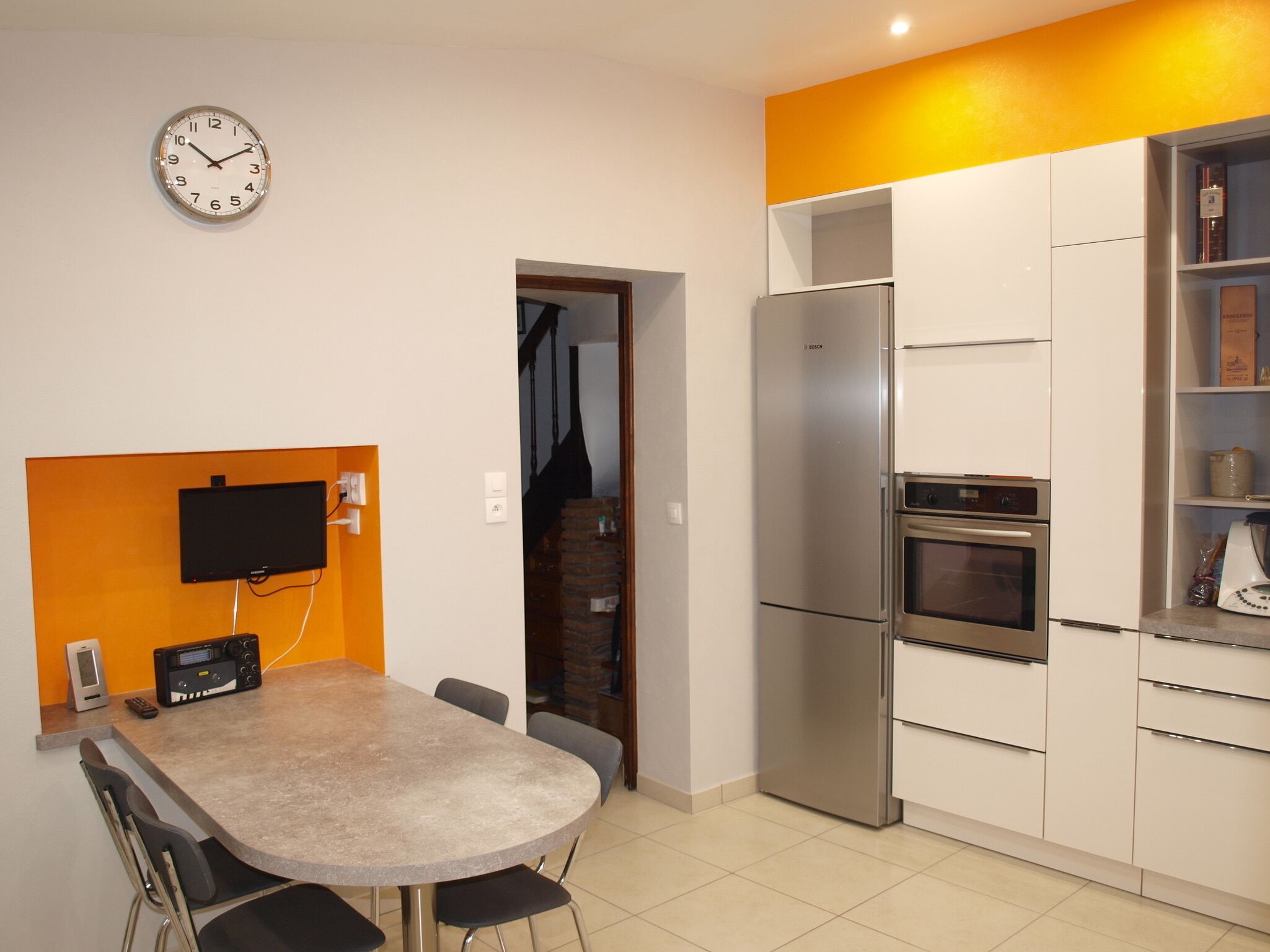 A modern kitchen with a built-in refrigerator, oven, and a flat-screen TV mounted on an orange wall.