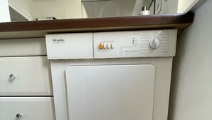 A compact, white, front-loading washing machine with a control panel featuring buttons and a dial, situated in a corner of a room with a tiled floor and a wooden countertop above.