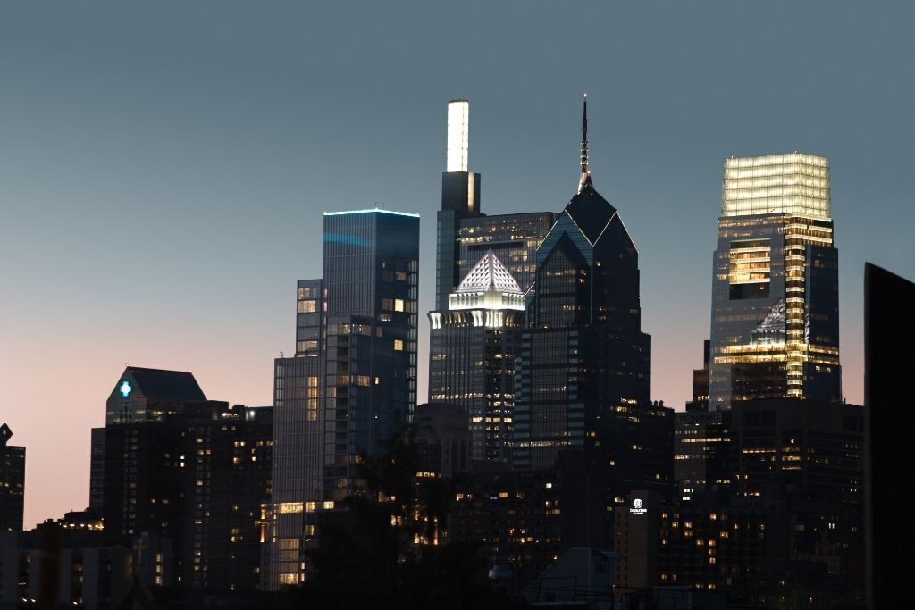A city skyline at dusk with illuminated buildings, including a prominent building with a pointed roof and a tall skyscraper with a spire.