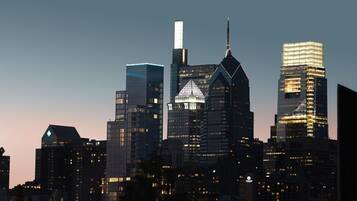 A city skyline at dusk with illuminated buildings, including a prominent building with a pointed roof and a tall skyscraper with a spire.