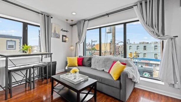 A modern living room with a grey sofa, wooden flooring, a dining table, and large windows offering a city view. - Brand New Modern Home /w Rooftop Downtown Skyview (Philadelphia)