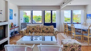 Ein geräumiges Wohnzimmer mit einer Couch, einem Couchtisch und Sesseln. Es gibt einen Kamin, einen Esstisch mit Stühlen und große Fenster mit Blick auf Grünflächen.