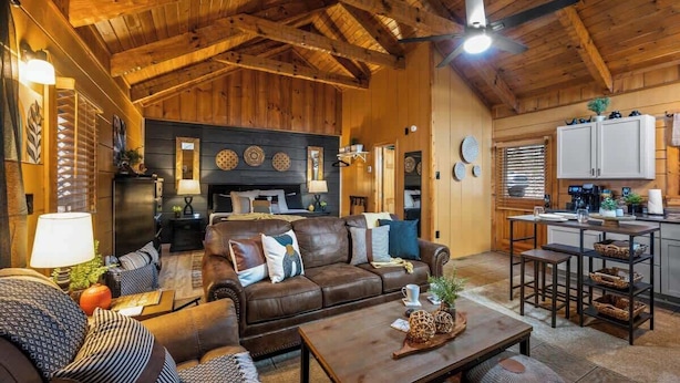 L’intérieur chaleureux d’un chalet avec un plafond en bois, un salon doté d’un canapé en cuir marron, un coin repas avec une table en bois et une cuisine avec des meubles blancs.