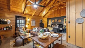 L’intérieur d’un chalet confortable, avec une salle à manger, un ensemble de canapés, un lit et un ventilateur de plafond.