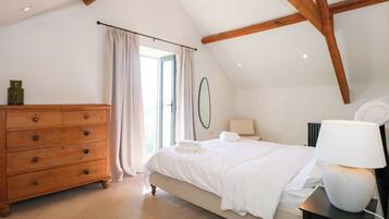 Une chambre à coucher avec une commode en bois, un lit recouvert de draps blancs, une lampe de chevet et une vue sur l’extérieur par une fenêtre ornée de rideaux.