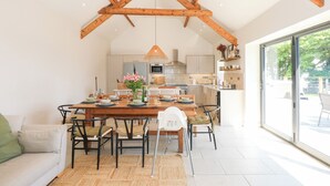 A spacious dining area with a wooden table and chairs, a kitchen in the background, and a large sliding glass door leading to an outdoor area. - Ysgubor Maen Y Bardd (Conwy)