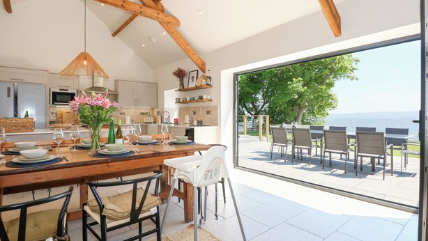 A modern kitchen with wooden beams, a dining area with a view, and a patio with outdoor seating. - Ysgubor Maen Y Bardd (Conwy)
