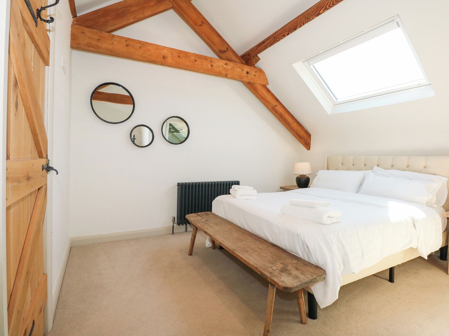 A bedroom with a sloped ceiling, wooden beams, a bed with white linens, a bench, a radiator, and three wall-mounted mirrors.