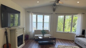 Un salón con chimenea, televisor, sofá, mesa de centro y ventanas amplias con vistas al agua.