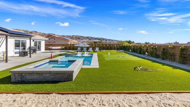 A modern outdoor pool area with a rectangular pool, a grassy lawn, and a sandy beach area. - Salida Del Sol by Rather Be Properties (Desert Hot Springs)