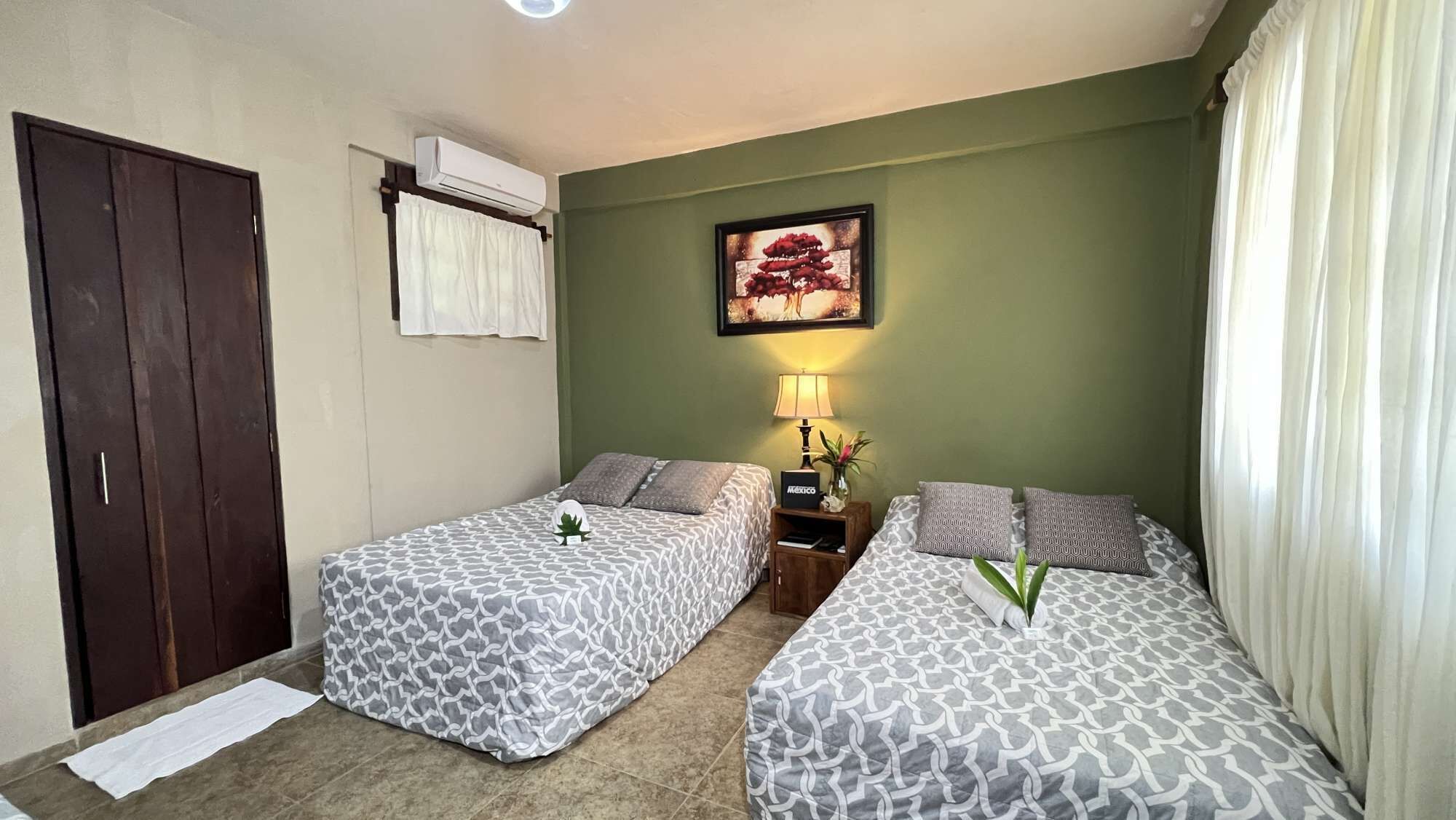 A hotel room with two beds, a wardrobe, an air conditioning unit, a framed picture, a nightstand with a lamp, and a window with curtains.
