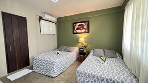 A hotel room with two beds, a wardrobe, an air conditioning unit, a framed picture, a nightstand with a lamp, and a window with curtains.