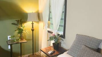 Una habitación con una cama, una mesita de noche con lámpara, una planta en maceta y un ventanal con cortinas.