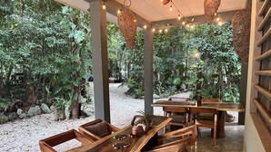 An outdoor wooden dining area with tables and chairs, surrounded by lush greenery and string lights.